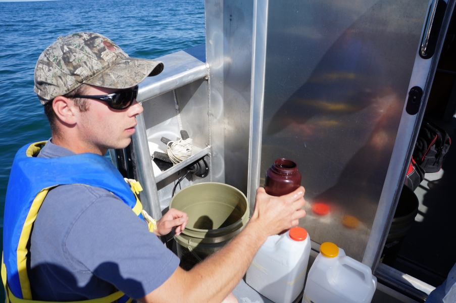 A person on a boat holds up a brown bottle. There is a bucket and two jugs nearby.