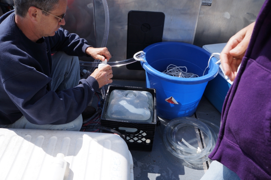 A person on a boat opens a bottle. They sit next to a cube container.