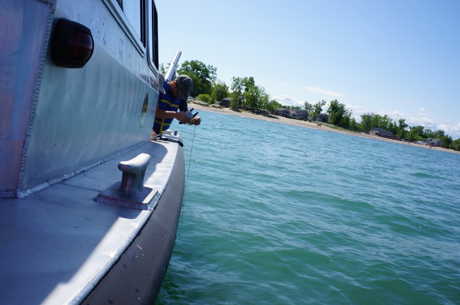 Looking along the side of a boat near a beach, someone is leaning over the side with a spool of rope.