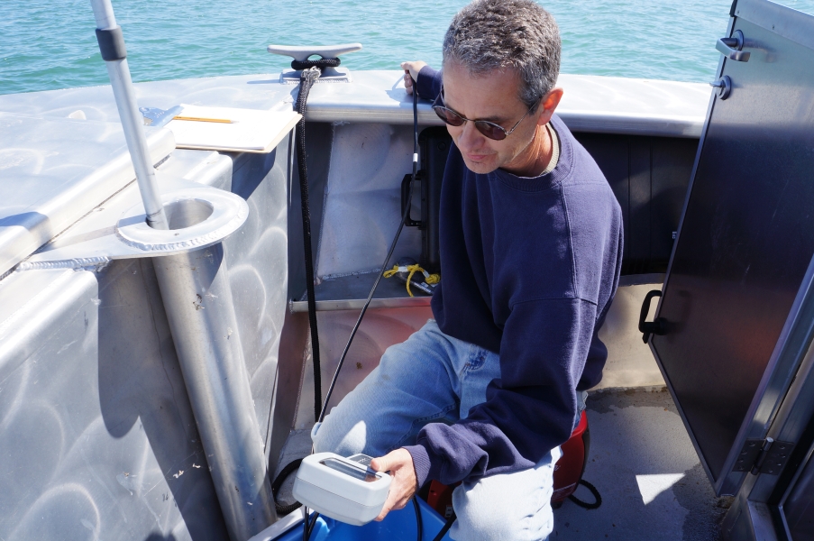 A person sits in the front of a boat, holding a small instrument with a cable going over the side of the boat.