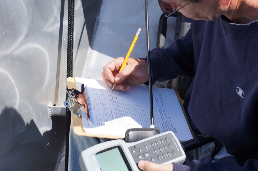 A person on a boat holding an instrument and recording the numbers on a data sheet on a clipboard