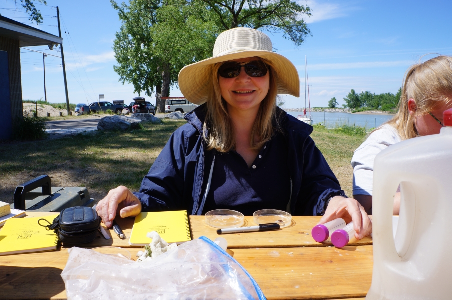 A person in a hat and sunglasses sits at a picnic table with petri dishes and small bottles.