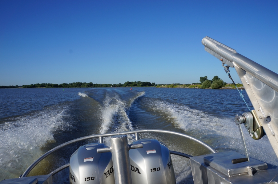 The back of a boat with two engines and a davit. The boat is throwing wake as it goes.