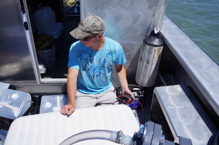 A person on the back deck of a boat sits among cube containers and a cooler. They are flipping a switch.