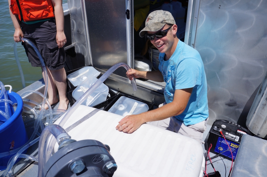 A person sits in the back of a boat, holding the end of a tube near a cube container. A second person is holding the other end in the water.