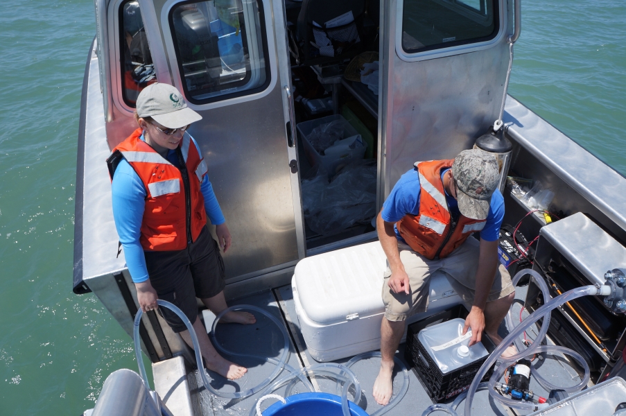 Two people on the back of a boat. One holds a tube over the side of the boat which loops across the deck, while a second opens up a cube container.