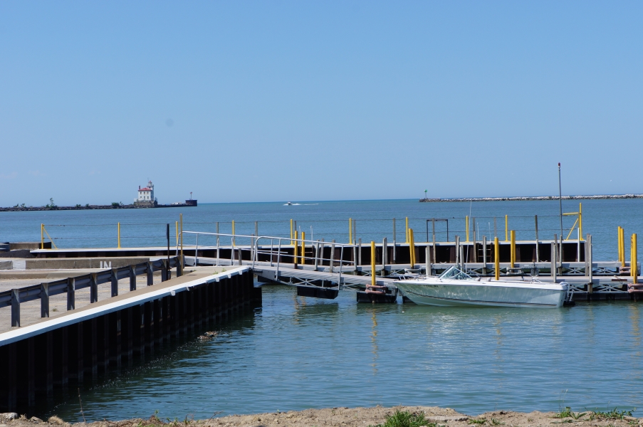 A small boat sitting in the water at a dock. In the far distance there is a pier with a two story building with a light house at the top.