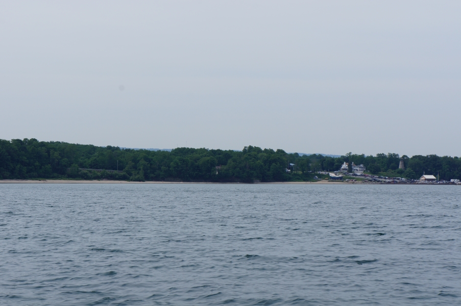 A parking lot with many cars by the water. There is a large house, a short stone lighthouse, and a wooden ship on land nearby.