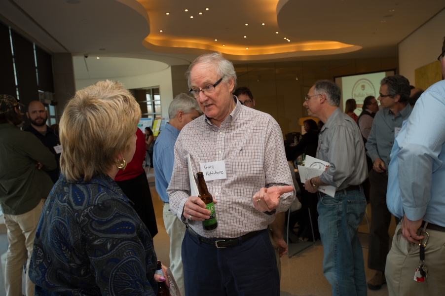 A person whose name tag reads "Gary Pettibone" talks with another person. There are many guests standing socializing behind them.