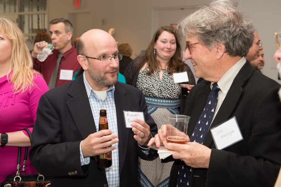 A person whose name tag reads "Russell" speaks with "John Montague." Several people are socializing behind them.