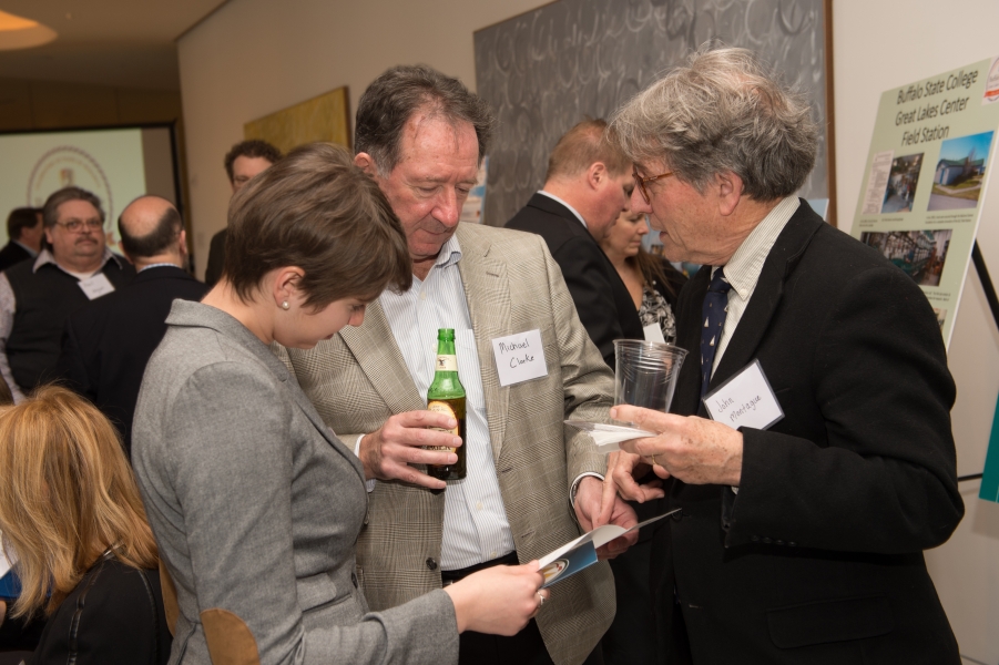 Three people chat while they look at a program pamphlet. Two of their nametags says "Michael Clarke" and "John Montague." Other people stand behind them