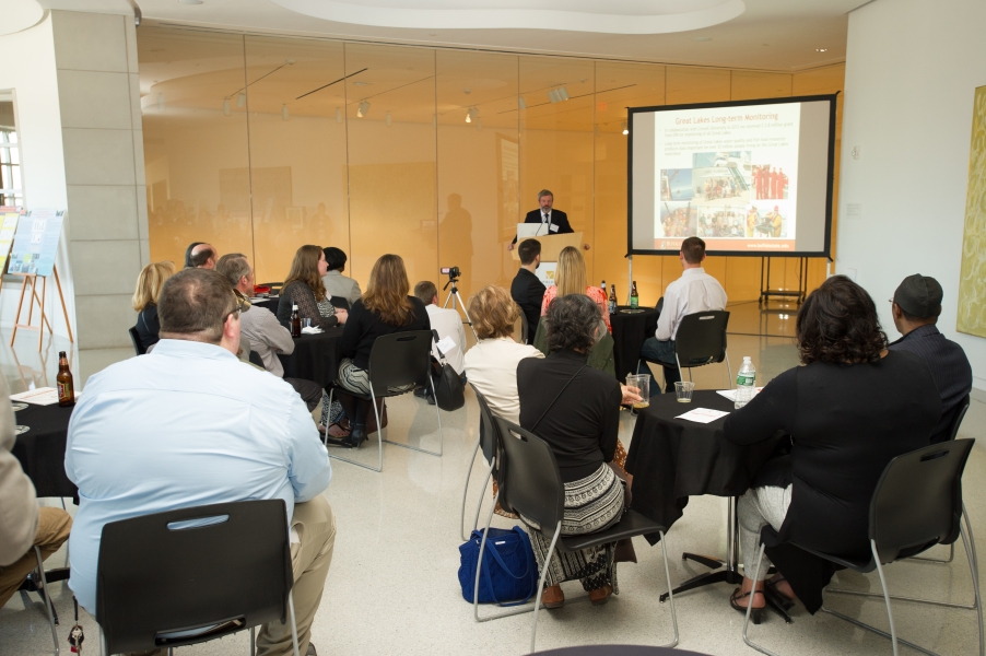 Guests watch as someone presents at a podium. A projection screen is next to the speaker.