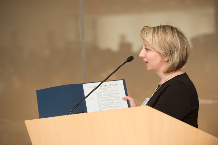 A person at a podium holds up a blue folder with a paper inside that says City of Buffalo