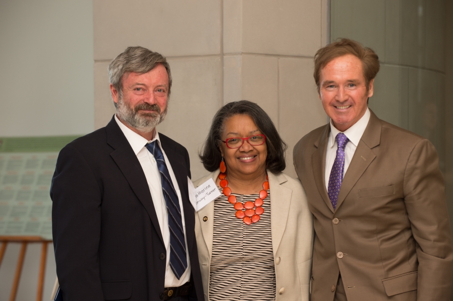 two people in suits flank a person wearing a blouse, blazer, and chunky necklace to pose for a photo