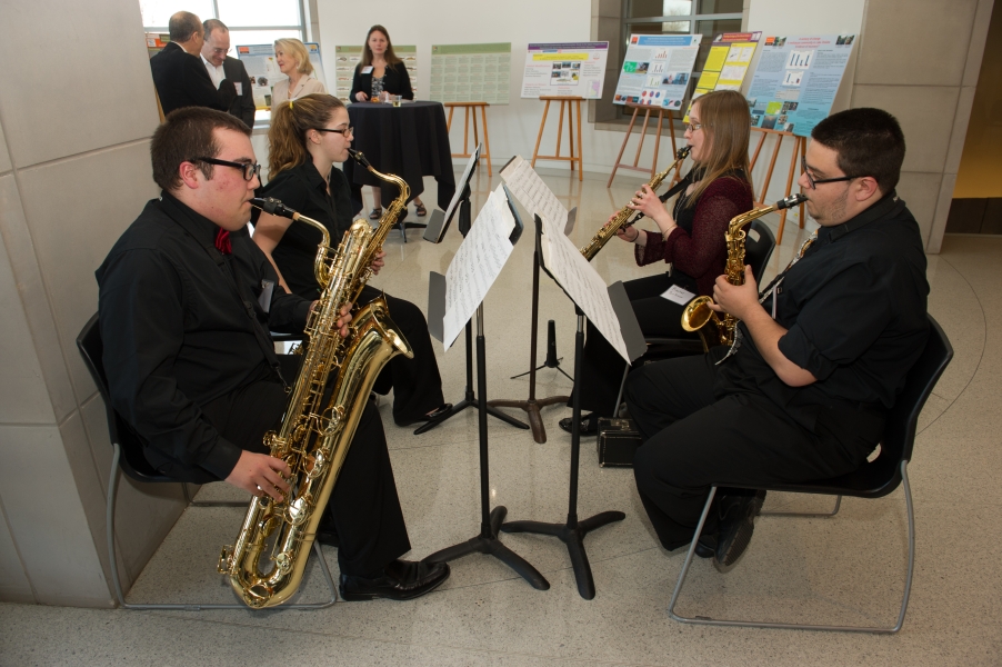 four people playing different types of saxophone sit facing each other with music stands. There are posters on easels and people in the background.