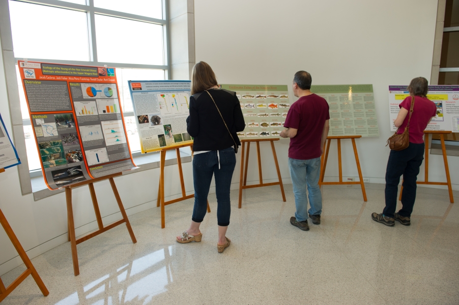 Three people stand before several easels with academic posters against the wall of a curved room with windows