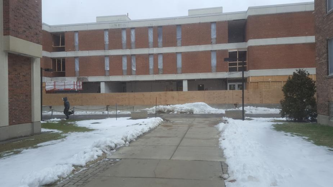 A construction site in winter. The windows on the second and third floor have been removed and covered with plastic. The first floor windows and walls have been removed to show the interior of the building. A wooden fence blocks the site from the walkway.