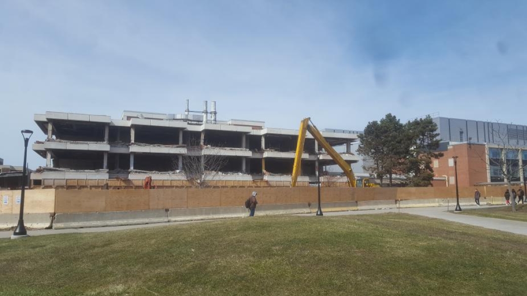 A construction site. The walls have been removed from this three-story academic building. Wooden fences surround the building. An excavator is behind the fence.