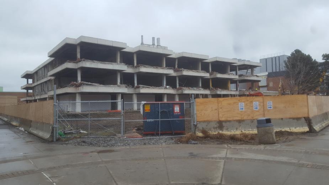 A construction site. The walls have been removed from this three-story academic building. Wooden fences surround the building. A Dumpster sits by a chain-link portion of the fence.