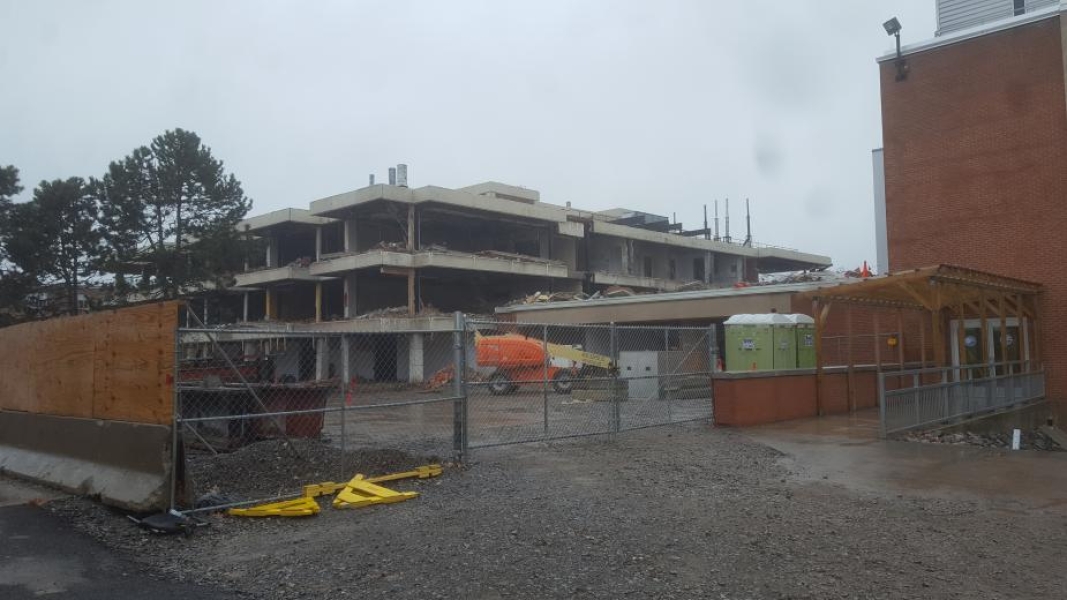 A construction site. The walls have been removed from this three-story academic building. Fences surround the building. A cherry picker is parked near some portapotties.