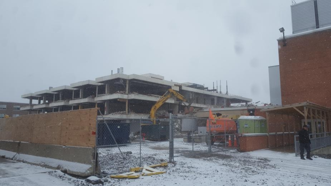 A construction site, where an excavator with a grapple is demolishing part of the building. The walls have been removed from this three-storey academic building. Fences surround the site, and there is snow on the ground.