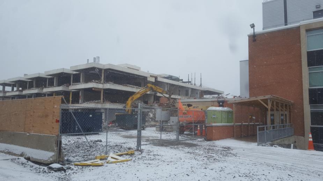 A construction site, where an excavator with a grapple is demolishing part of the building. The walls have been removed from this three-storey academic building. Fences surround the site, and there is snow on the ground.