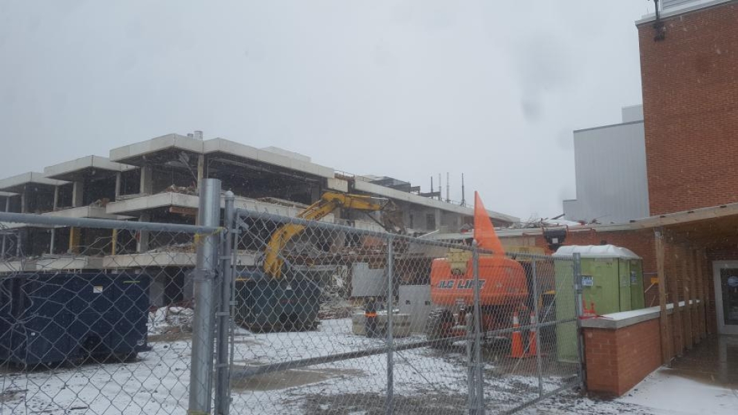 A construction site, where an excavator with a grapple is demolishing part of the building. The walls have been removed from this three-storey academic building. Fences surround the site, and there is snow on the ground.