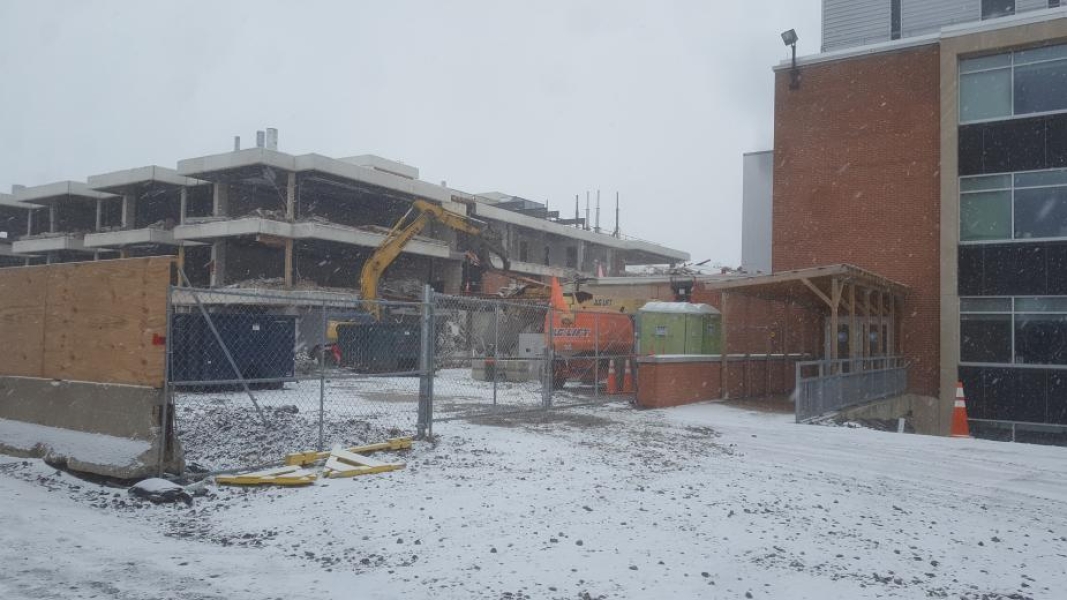 A construction site, where an excavator with a grapple is demolishing part of the building. The walls have been removed from this three-storey academic building. Fences surround the site, and there is snow on the ground.