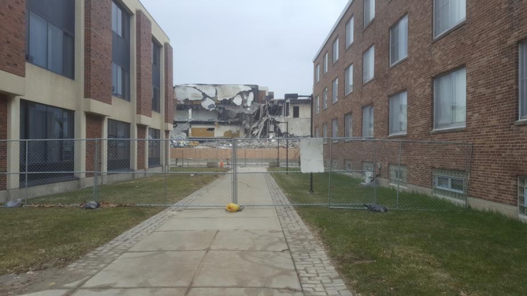 A view between two academic buildings. A construction site is blocked off with multiple fences, and a building is being demolished. Parts of the building hang down onto the floors below.