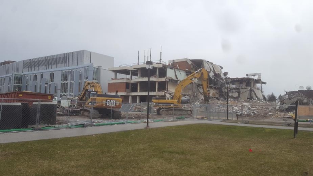 A construction site with a partially demolished building. Two excavators work in the foreground, and part of the roof of the building droops down onto the floors below.