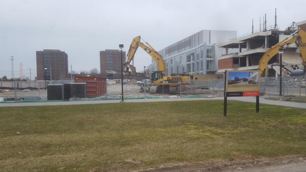 An excavator manipulates a steel beam as a second one works at a construction site. A sign outside of the chain-link fence describes the construction project.