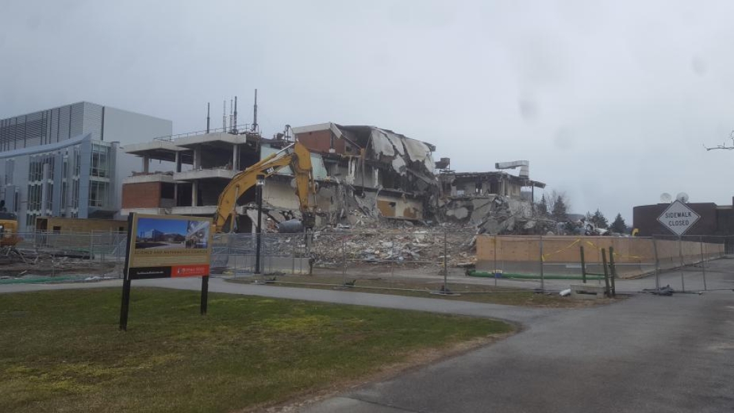 An excavator working through piles of rubble by a partially demolished building. A sign outside of the chain-link fence describes the construction project.