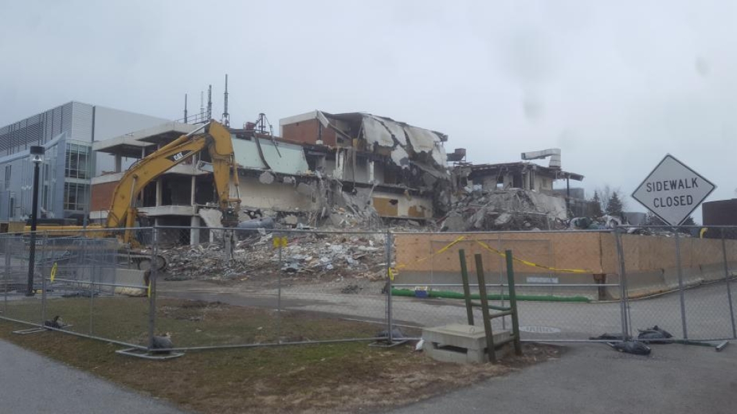 A construction site with a partially demolished building. An excavator work in the foreground, and part of the roof of the building droops down onto the floors below.