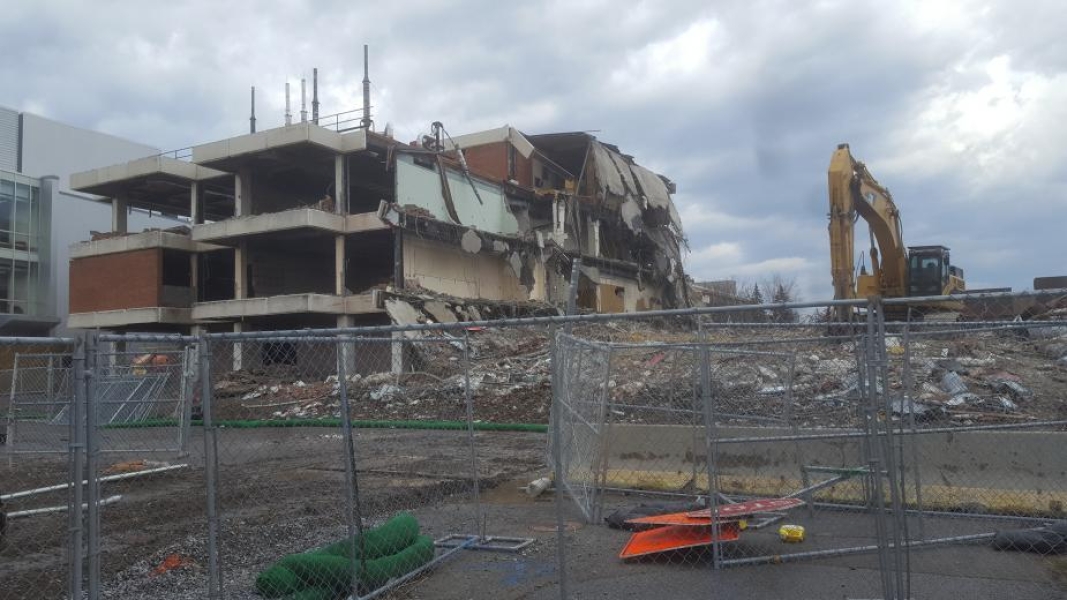 A construction site with a partially demolished building. The roof droops over the lower levels. An excavator is working in the rubble.