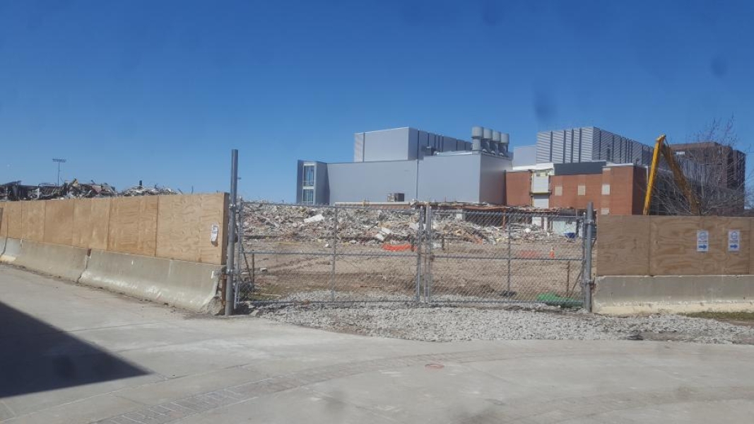 A field of debris next to a grey building and a brick building. The construction area is surrounded by a wooden fence that is interrupted by a chain-link fence through which we can see the debris.