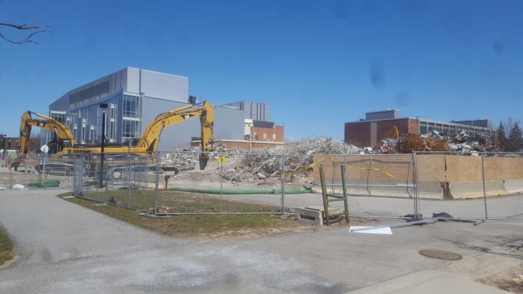 A field of debris next to a grey building and a brick building. The construction area is surrounded by chain-link fencing. Two excavators are parked in the construction zone.