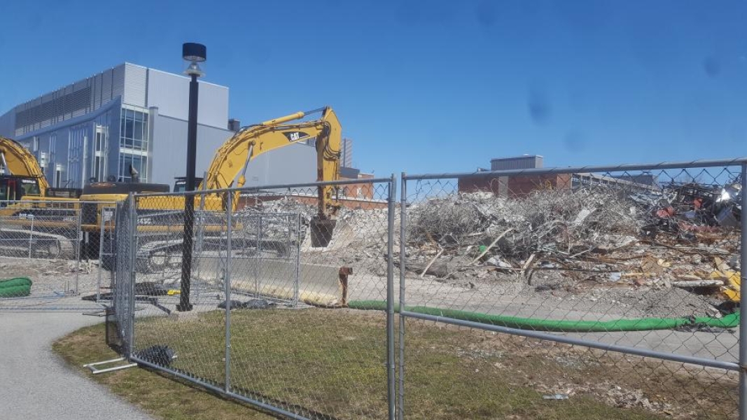 A field of debris next to a grey building and a brick building. The construction area is surrounded by chain-link fencing. Two excavators are parked in the construction zone.