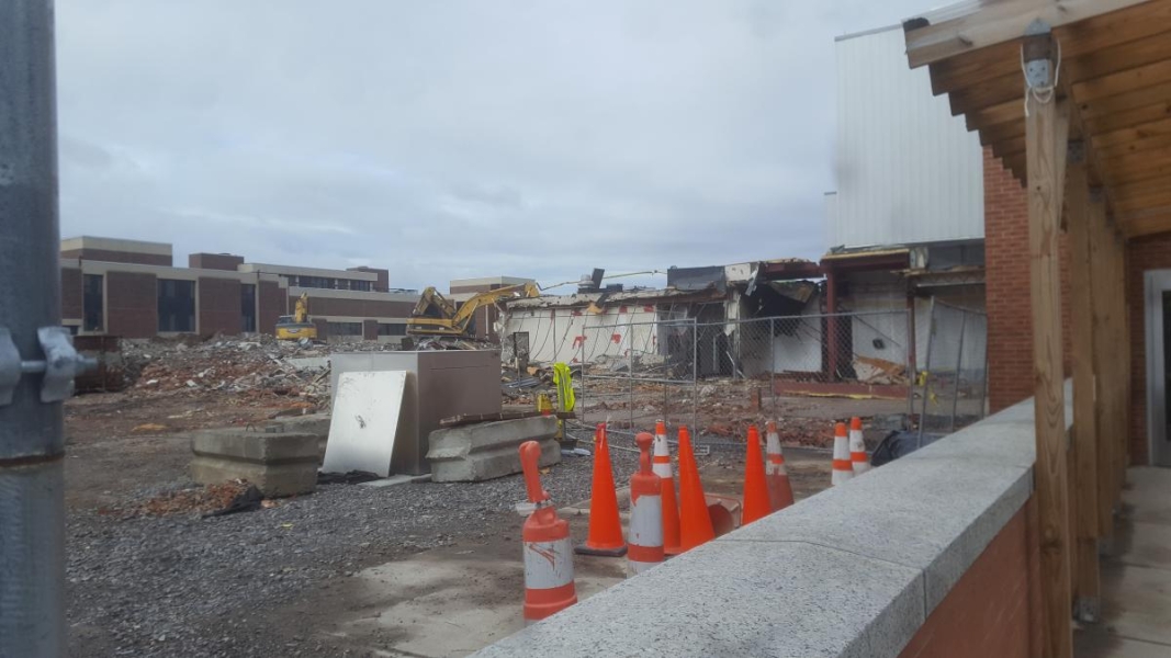 A construction site with a single-level part of a demolished building and some safety cones in the foreground, and piles of rubble and excavators in the background.