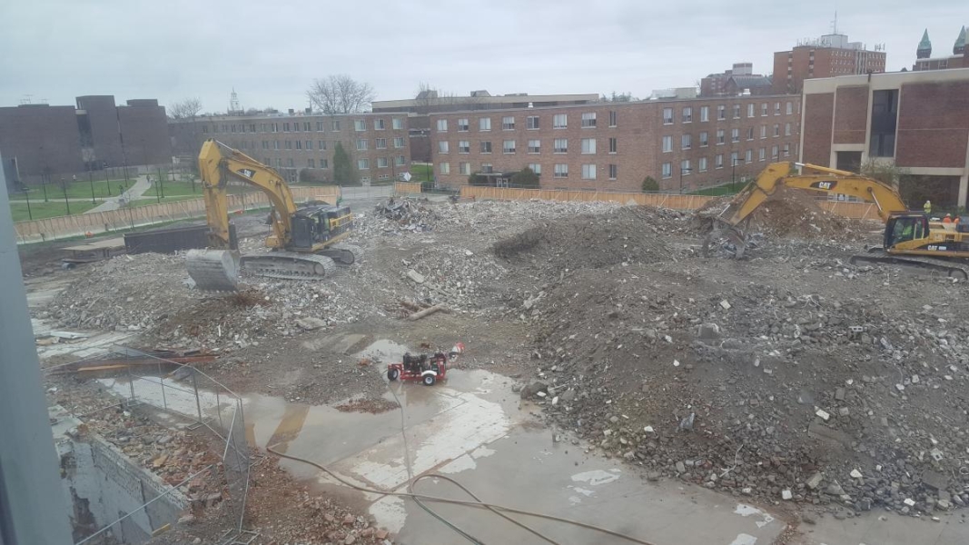 A field of rubble in a construction site, but there is some ground showing around the piles of rubble. Two excavators work to move and sort rubble. There is a generator on the clear ground.