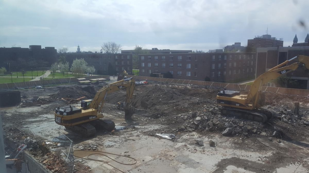 Two excavators in a construction site. One is on flat concrete and the other is on a pile of rubble on the concrete. Beyond the excavators, the ground is being excavated.