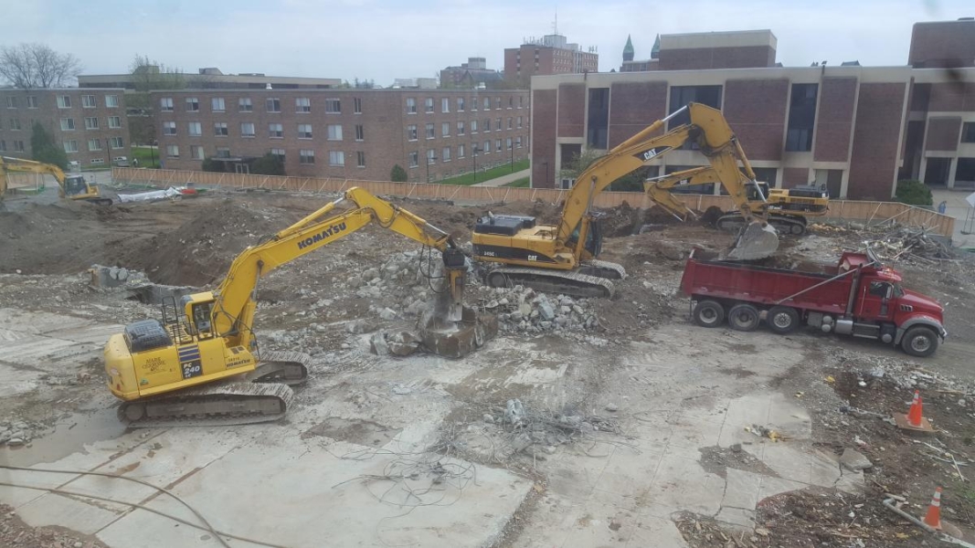 A construction site. One excavator is loading rubble into a dump truck. Another is using a jack hammer arm to break apart huge pieces of concrete. Two other excavators dig in the background.