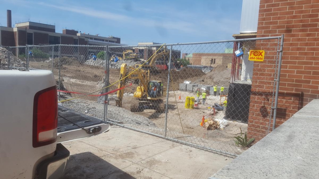 A construction site next to a building. Construction workers walk on the foundation near pallets of cinder blocks. A small excavator works in the foreground while other larger ones remove rubble in the background.