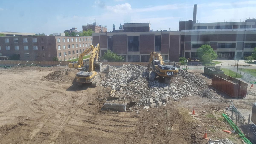 A construction site with concrete rubble in one corner and bare dirt elsewhere. Three excavators work on the rubble.