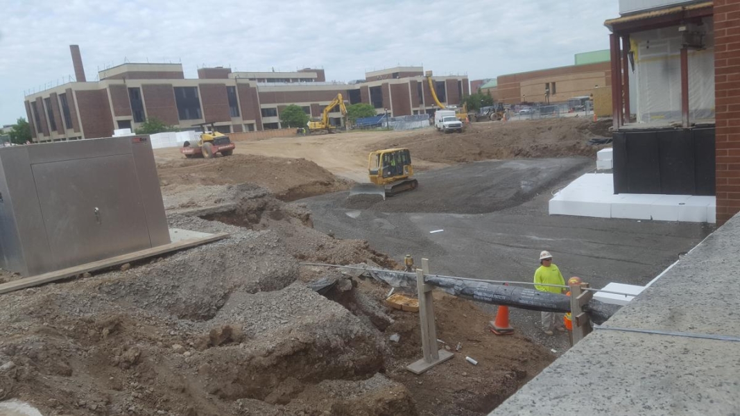 A small bulldozer spreads gravel over a foundation. Two workers inspect the foundation of the existing building in the foreground.