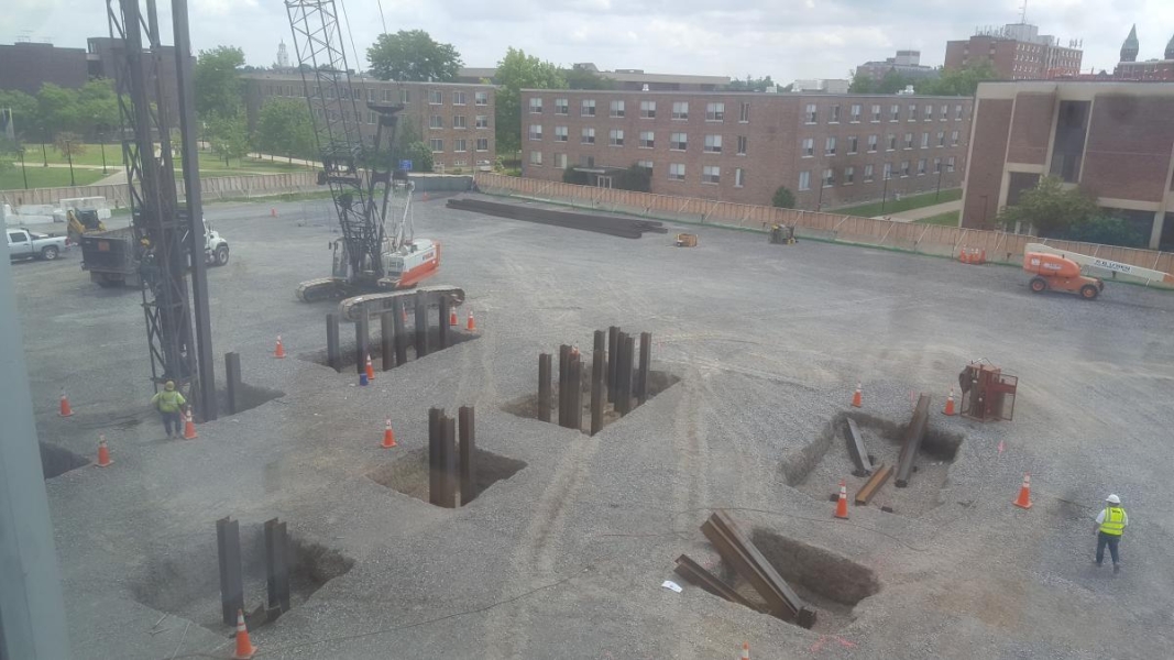 A crane pounding steel beams into sqaure holes in the ground. There are eight holes and each have multiple beams driven into them.