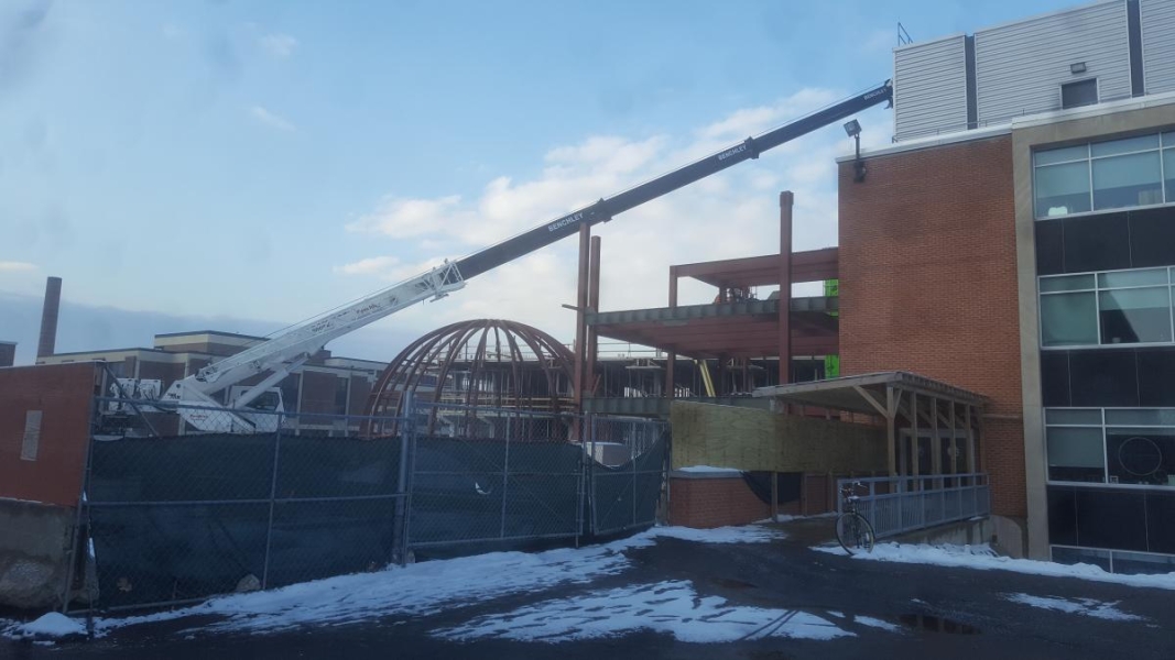 A crane reaches to the top floor of a partially constructed building. There are steel beams, including steel beams in a dome shape.