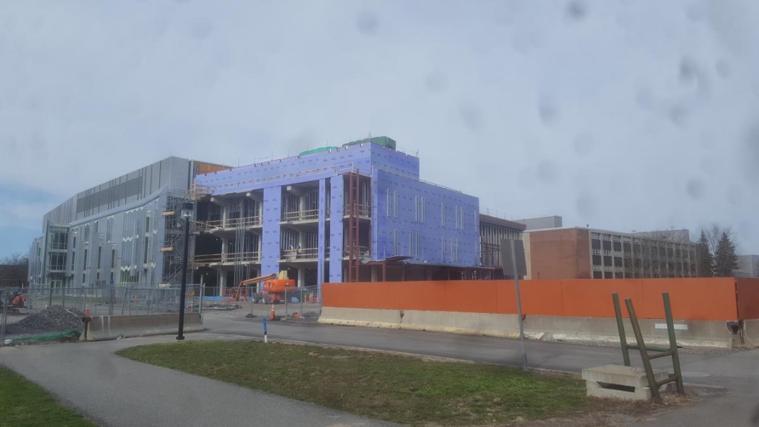 A partially constructed building. The existing part of the building is metal with many windows. The new part of the building is partially enclosed with walls, but other places are open where large windows later be placed. The walls inside are framed but not built yet.