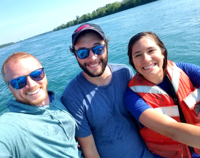 Three smiling people stand in front of the water. Two men wearing sunglasses and a woman in a life jacket