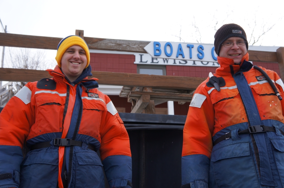 two people in flotation suits and knit caps stand in front of a building with a sign "Boats"