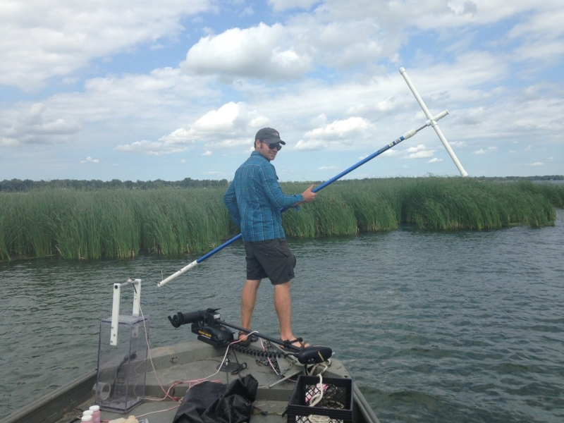 A person stands on the front of a boat holding a large t-shaped pole. They are near a wetland with emergent vegetation.